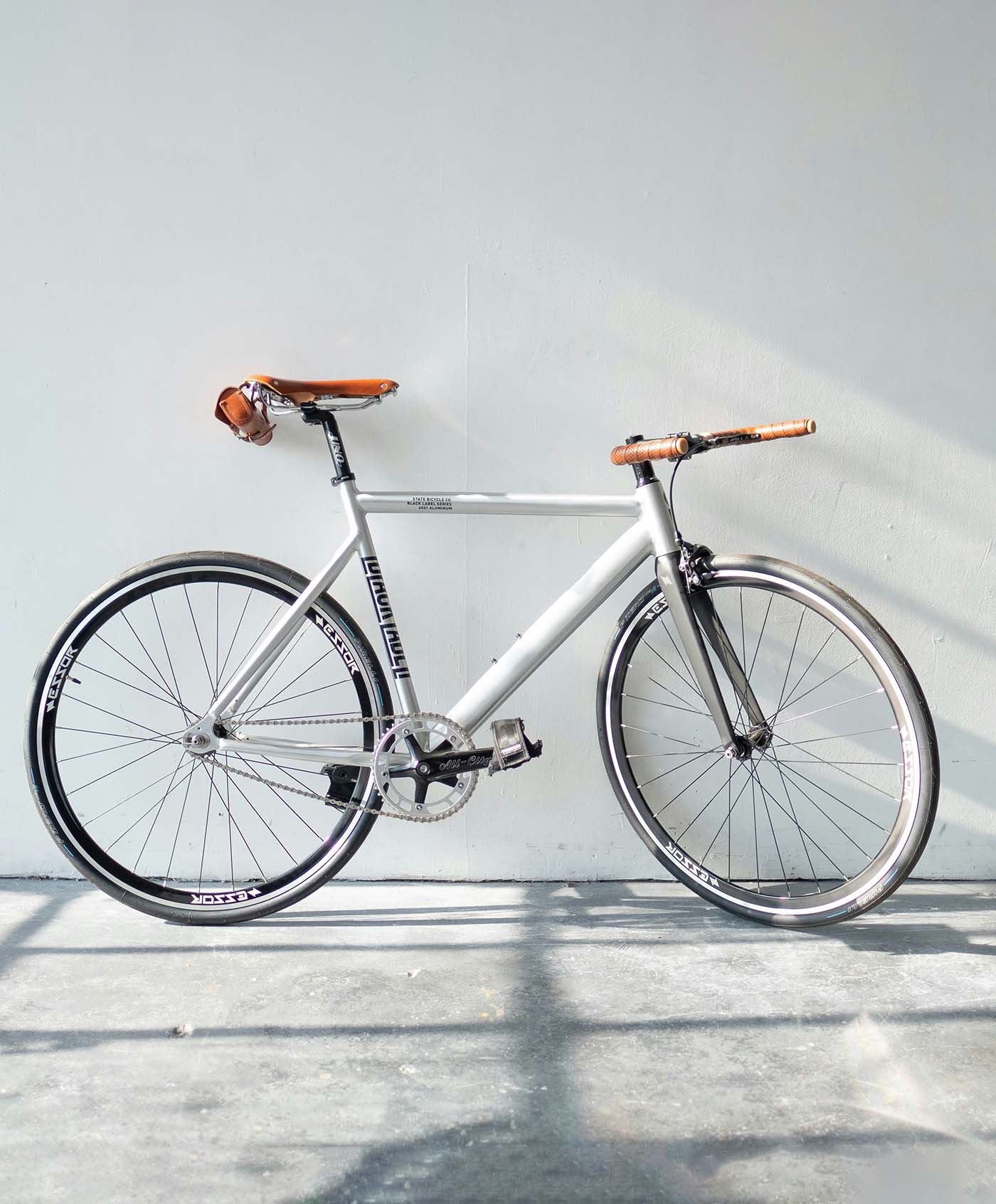 A white fixed gear bicycle with brown leather saddle and handlebars, leaning against a wall in direct sunlight.