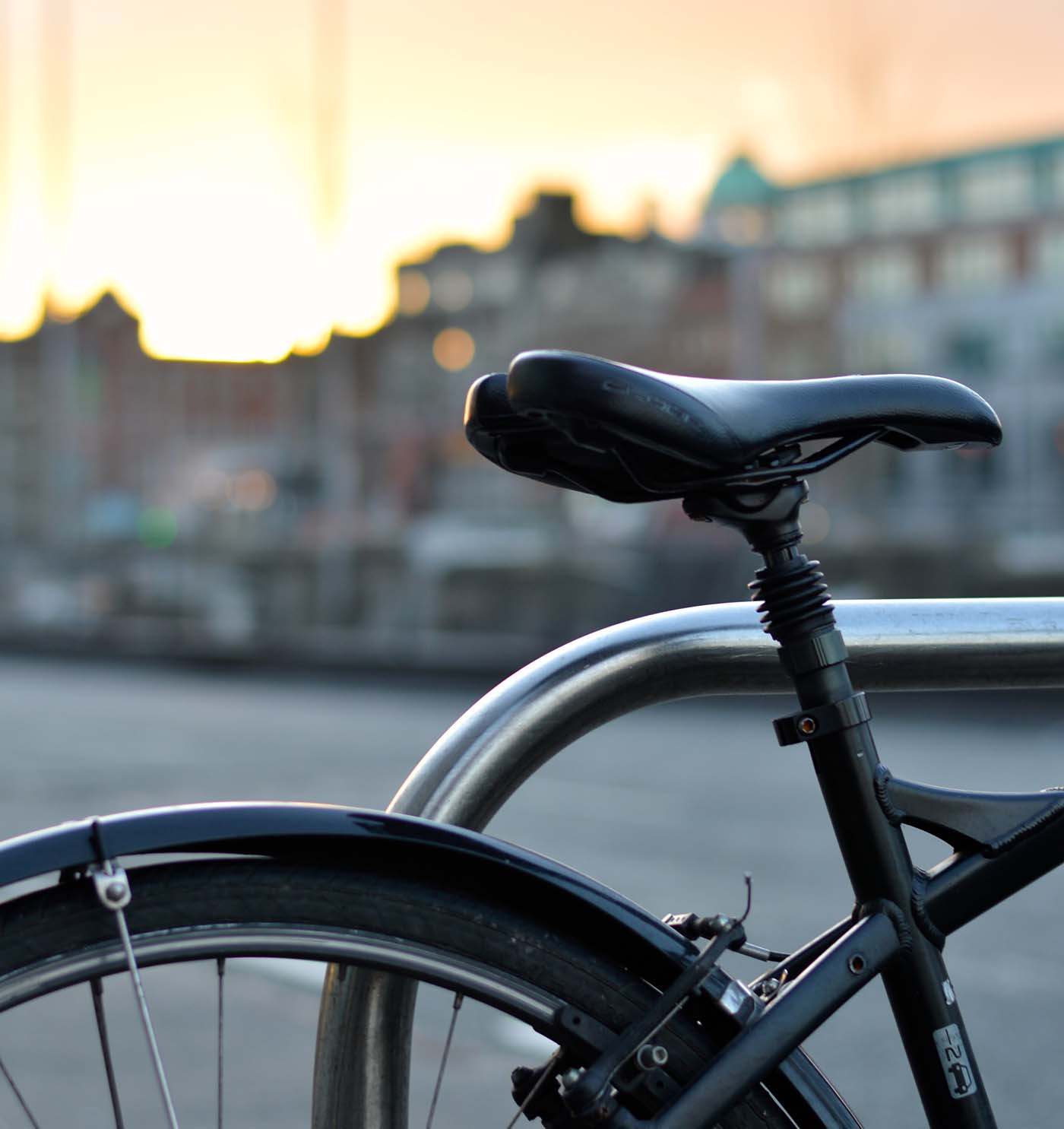 A close-up of a black bicycle seat and rear tire with a city skyline blurred in the background during sunset.